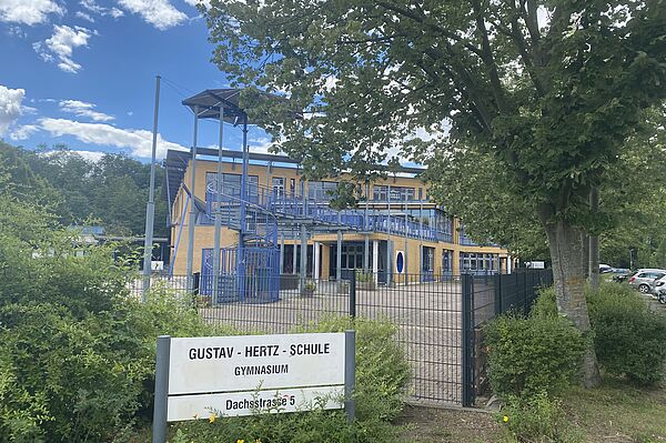 Die Gustav-Hertz-Schule verfügt über eine moderne Architektur mit blauen Geländern und einer markanten Wendeltreppe, die in das Obergeschoss führt. Große Fensterfronten ermöglichen einen guten Lichteinfall in die Innenräume. Das Schulgebäude ist von einem gepflegten Schulhof umgeben. Der Eingangsbereich wird durch ein Schild mit der Bezeichnung der Schule klar gekennzeichnet.