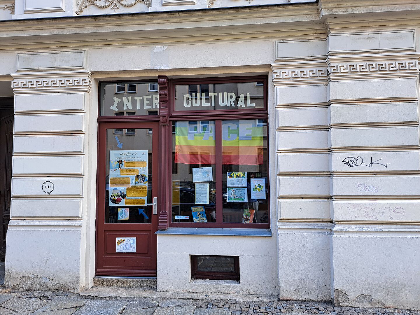  Das Bild zeigt die Fassade der "Bunten Büchothek", ein kulturelles Zentrum, erkennbar an den mit Plakaten und Informationen dekorierten Fenstern. Die Eingangstür ist rot, und das Fenster daneben trägt die Aufschrift "INTERCULTURAL" in großen Lettern. Die Fassade des Gebäudes weist klassische Wandverzierungen in Weiß und Creme auf. Graffiti ist ebenfalls an den Wänden sichtbar.