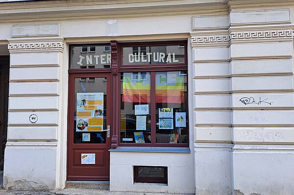  Das Bild zeigt die Fassade der "Bunten Büchothek", ein kulturelles Zentrum, erkennbar an den mit Plakaten und Informationen dekorierten Fenstern. Die Eingangstür ist rot, und das Fenster daneben trägt die Aufschrift "INTERCULTURAL" in großen Lettern. Die Fassade des Gebäudes weist klassische Wandverzierungen in Weiß und Creme auf. Graffiti ist ebenfalls an den Wänden sichtbar.