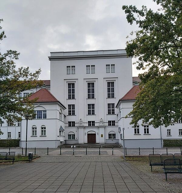 Das Bild zeigt die Fassade eines imposanten, weißen Gebäudes, das zur „Landesschule für Hör- und Sprachgeschädigtenwesen“ gehört. Das Gebäude hat eine schlichte, aber elegante Architektur mit symmetrischen Linien und hohen Fenstern. Der Mittelteil des Gebäudes ist dominant und hat vertikale Fenster, die die Höhe betonen, während die Seitenflügel des Baus mit roten Ziegeldächern bedeckt sind. Der Eingangsbereich ist verziert, und das Hauptportal ist umrahmt von Details im Stil der klassischen Architektur.