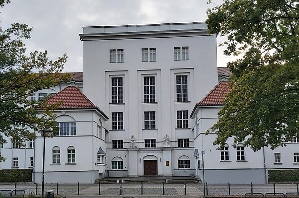 Das Bild zeigt die Fassade eines imposanten, weißen Gebäudes, das zur „Landesschule für Hör- und Sprachgeschädigtenwesen“ gehört. Das Gebäude hat eine schlichte, aber elegante Architektur mit symmetrischen Linien und hohen Fenstern. Der Mittelteil des Gebäudes ist dominant und hat vertikale Fenster, die die Höhe betonen, während die Seitenflügel des Baus mit roten Ziegeldächern bedeckt sind. Der Eingangsbereich ist verziert, und das Hauptportal ist umrahmt von Details im Stil der klassischen Architektur.