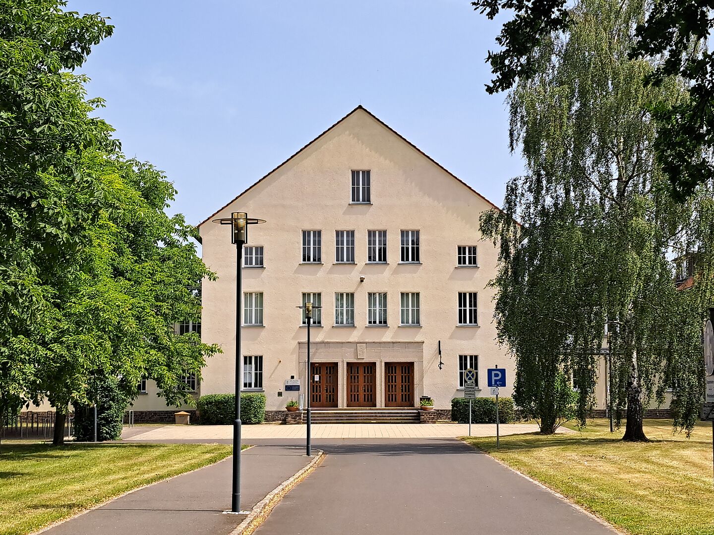 Die Außenansicht der Ruth-Pfau-Schule zeigt ein dreistöckiges Gebäude mit einer beigefarbenen Fassade und einem Satteldach. In der Mitte des Gebäudes befindet sich ein Eingang mit drei großen Holztüren und einer kleinen Treppe. Vor dem Eingang stehen zwei Laternen. Der Weg zum Eingang ist asphaltiert und von grünen Rasenflächen sowie Bäumen und Sträuchern gesäumt. Auf der rechten Seite des Gebäudes ist ein Behindertenparkplatz mit einem entsprechenden Schild zu sehen. Der Himmel ist klar und blau.