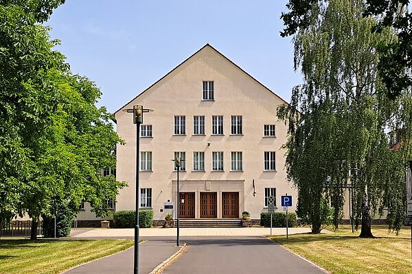 Die Außenansicht der Ruth-Pfau-Schule zeigt ein dreistöckiges Gebäude mit einer beigefarbenen Fassade und einem Satteldach. In der Mitte des Gebäudes befindet sich ein Eingang mit drei großen Holztüren und einer kleinen Treppe. Vor dem Eingang stehen zwei Laternen. Der Weg zum Eingang ist asphaltiert und von grünen Rasenflächen sowie Bäumen und Sträuchern gesäumt. Auf der rechten Seite des Gebäudes ist ein Behindertenparkplatz mit einem entsprechenden Schild zu sehen. Der Himmel ist klar und blau.