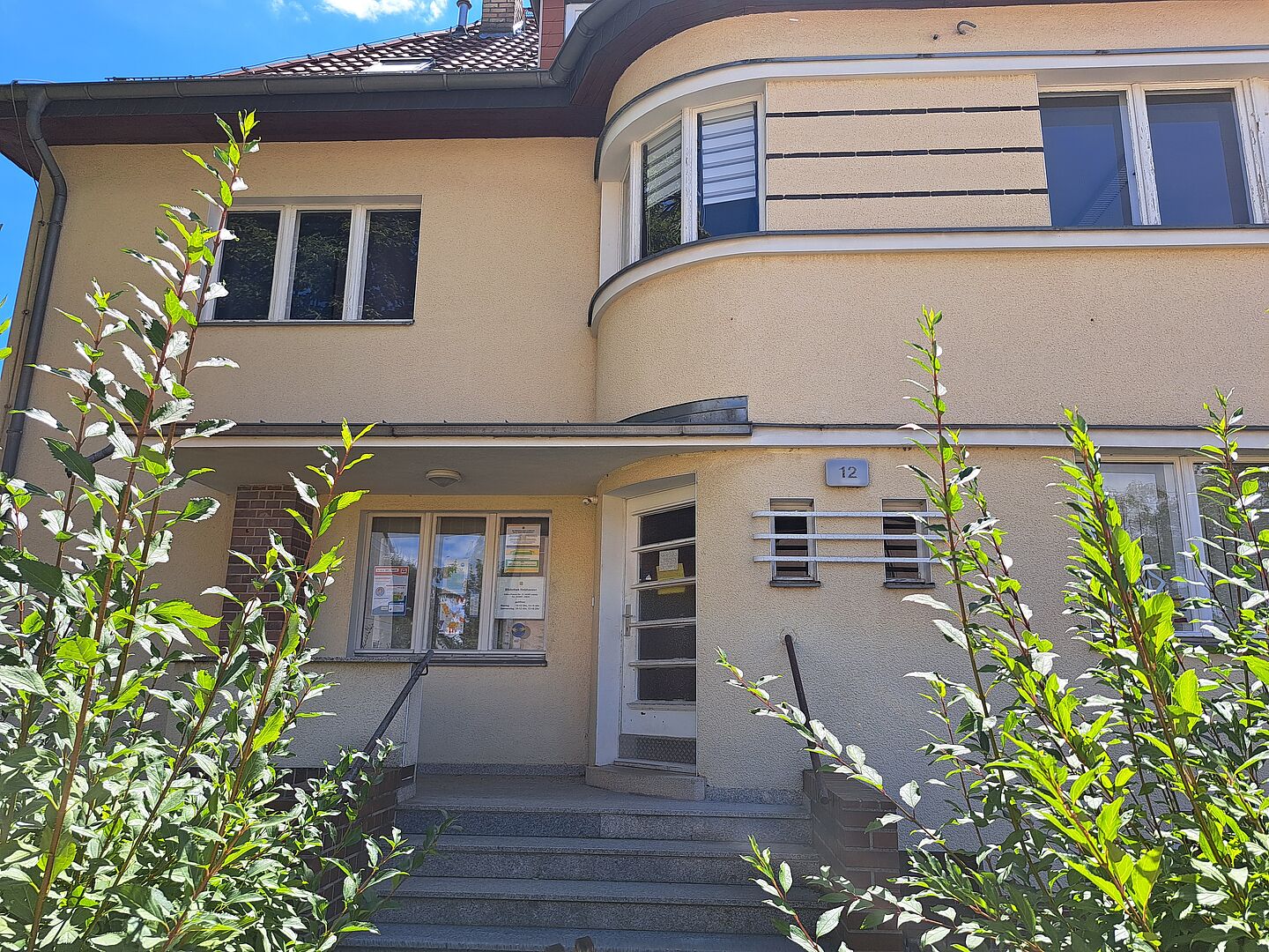 Das Bild zeigt den Eingang der Stadtteilbibliothek Holzhausen. Das Gebäude hat eine helle Fassade und ist zweistöckig. Der Eingang besteht aus einer Glastür, die über eine kleine Treppe erreichbar ist. Links neben der Tür ist ein Fenster mit Informationsplakaten. Rechts neben der Tür ist die Hausnummer 12 angebracht. Einige grüne Büsche sind im Vordergrund zu sehen.
