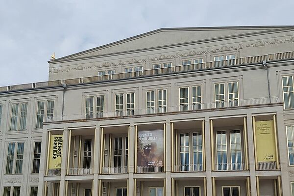 Das Bild zeigt das Gebäude der Oper Leipzig. Die Architektur ist klassisch-modern und repräsentativ. Die Fassade des Opernhauses ist symmetrisch und in einem hellen Ton gehalten. Auffällig sind die großen Fenster und die Säulen im Eingangsbereich, die den Haupteingang mit seinem imposanten Vorbau betonen.