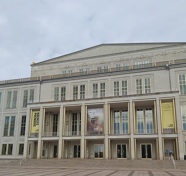 Das Bild zeigt das Gebäude der Oper Leipzig. Die Architektur ist klassisch-modern und repräsentativ. Die Fassade des Opernhauses ist symmetrisch und in einem hellen Ton gehalten. Auffällig sind die großen Fenster und die Säulen im Eingangsbereich, die den Haupteingang mit seinem imposanten Vorbau betonen.