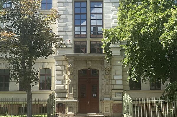 Das Bild zeigt die Fassade eines großen, historischen Gebäudes mit der Inschrift „KNABEN“ oben. Das Gebäude hat einen klassischen Architekturstil mit großen Fenstern und aufwändigem Steinwerk um den Eingang herum. Auf beiden Seiten des Eingangs stehen Bäume, und eine kleine Treppe führt zur Haupttür.