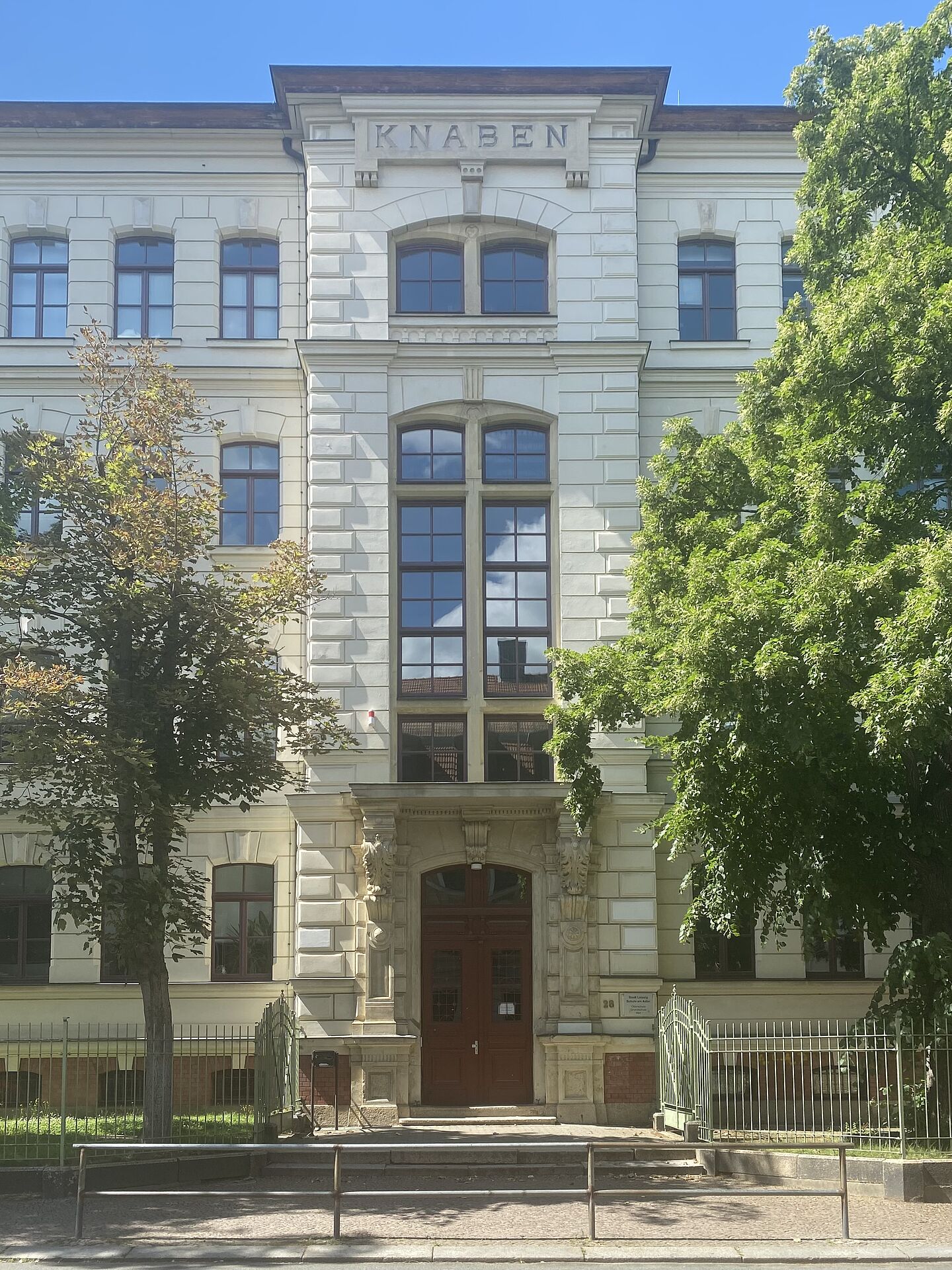 Das Bild zeigt die Fassade eines großen, historischen Gebäudes mit der Inschrift „KNABEN“ oben. Das Gebäude hat einen klassischen Architekturstil mit großen Fenstern und aufwändigem Steinwerk um den Eingang herum. Auf beiden Seiten des Eingangs stehen Bäume, und eine kleine Treppe führt zur Haupttür.