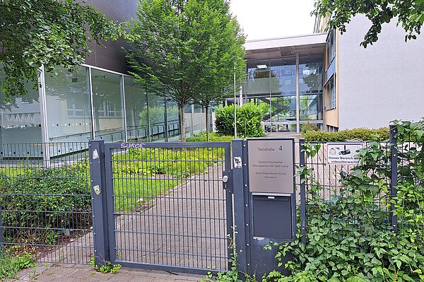 Das Bild zeigt den Eingangsbereich der Anton-Philipp-Reclam-Schule, ein modernes Schulgebäude mit großen Glasfassaden und abwechselnd dunklen und hellen Bauelementen. An einem Metalltor im Vordergrund steht die Adresse "Tarotstraße 4". Das Schulgelände ist von grünen Bäumen und Büschen umgeben, und ein Schild informiert über die Videoüberwachung des Bereichs.