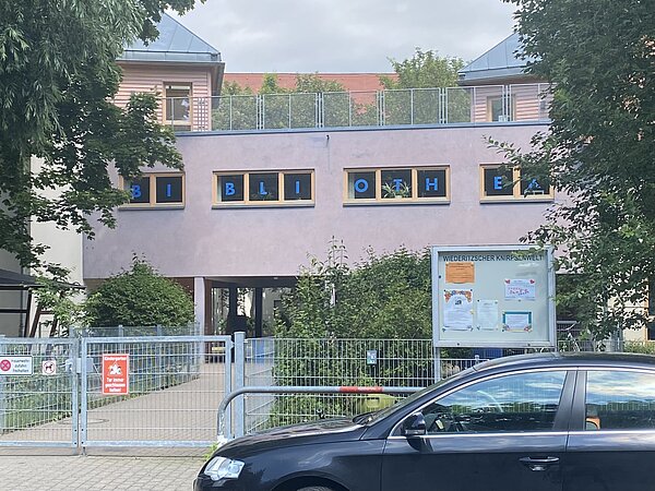 Leipziger Stadtteilbibliothek Wiederitzsch Das Bild zeigt die Außenansicht einer Bibliothek mit der Aufschrift "BIBLIOTHEK" in blauen Buchstaben auf den Fenstern im oberen Stockwerk. Die Fassade des Gebäudes ist hellrosa und das Dach ist blau. Vor dem Gebäude befindet sich ein Metalltor mit mehreren Schildern, darunter eines, das "Kindergarten" und ein weiteres, das "Tor immer geschlossen halten" besagt.