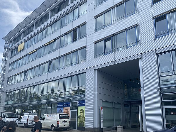Leipziger Stadtteilbibliothek Volkmarsdorf Das Bild zeigt ein modernes, mehrstöckiges Bürogebäude mit einer metallischen Fassade und zahlreichen großen Fenstern. Es hat mindestens fünf Stockwerke, und im Erdgeschoss gibt es einen überdachten Eingangsbereich.
