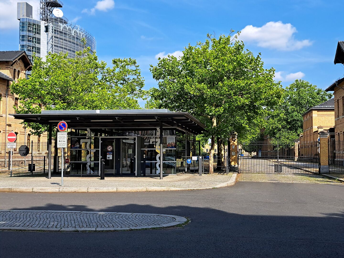 Das Bild zeigt die Zufahrt zum Gelände des Mitteldeutschen Rundfunks (MDR). Der Eingangsbereich ist durch ein modernes Pförtnerhaus  gekennzeichnet, das sich vor dem Tor des Geländes befindet. Hinter dem Eingang sind ältere Backsteingebäude sichtbar, die zum MDR-Komplex gehören. Reiche Grünflächen mit ausgewachsenen Bäumen umgeben den Eingangsbereich.