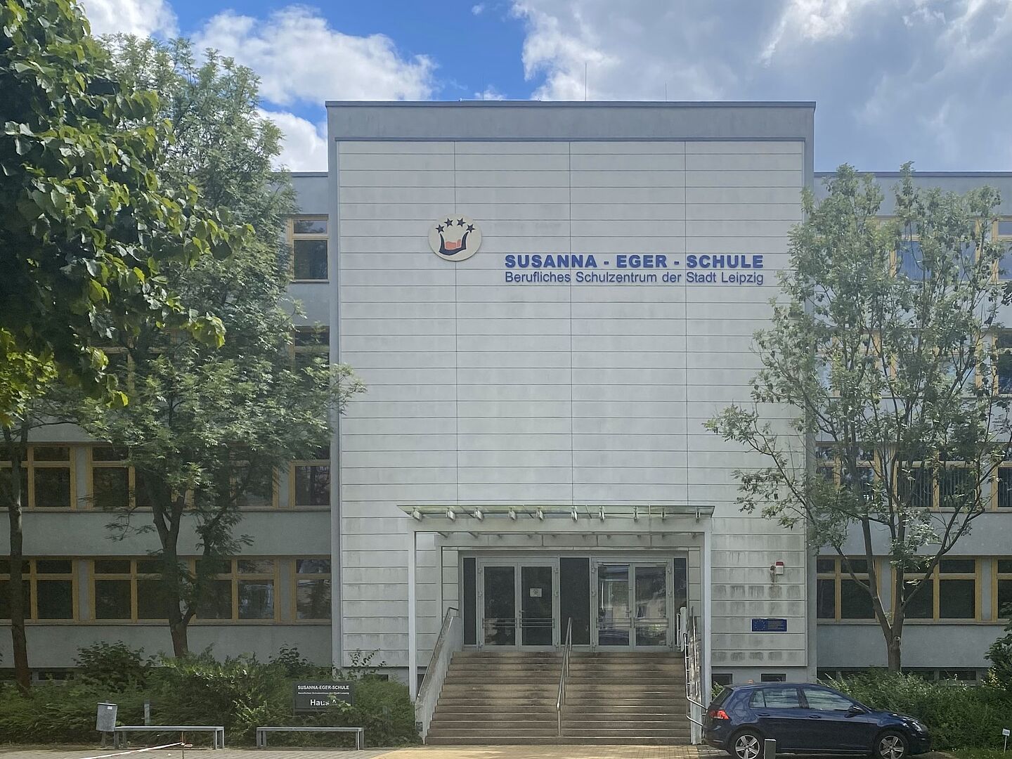 Das Bild zeigt die Susanna-Eger-Schule, ein Berufliches Schulzentrum der Stadt Leipzig. Das moderne Gebäude hat eine helle Fassade und ist mit dem Schullogo und dem Schriftzug deutlich gekennzeichnet. Vor dem Haupteingang befinden sich einige Stufen, die von Bäumen und Grünflächen umgeben sind. Die Susanna-Eger-Schule besitzt eine große Glastür am Haupteingang. Die Fenster des Gebäudes sind gleichmäßig angeordnet.