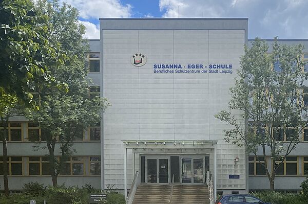 Das Bild zeigt die Susanna-Eger-Schule, ein Berufliches Schulzentrum der Stadt Leipzig. Das moderne Gebäude hat eine helle Fassade und ist mit dem Schullogo und dem Schriftzug deutlich gekennzeichnet. Vor dem Haupteingang befinden sich einige Stufen, die von Bäumen und Grünflächen umgeben sind. Die Susanna-Eger-Schule besitzt eine große Glastür am Haupteingang. Die Fenster des Gebäudes sind gleichmäßig angeordnet.