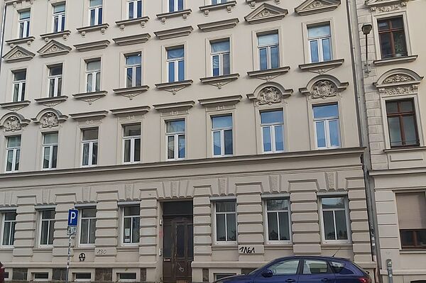 Das Bild zeigt die Fassade eines klassischen, mehrstöckigen Wohn- oder Bürogebäudes, das unter anderen das Institut für Kultur- und Universalgeschichte der Karl-Lamprecht-Gesellschaft e.V. beherbergt. Die Fassade ist in einem eleganten Stil gehalten, typisch für Gründerzeitbauten, mit klaren Linien und symmetrischen Fenstern. Über einigen Fenstern sind dekorative Giebel angebracht, die dem Gebäude zusätzliche architektonische Details verleihen. Das Erdgeschoss hebt sich durch größere Fenster und eine geradlinige Fassadengestaltung ab, die von horizontalen Bändern unterbrochen wird. Die Haustür ist schlicht, weist jedoch typische alte Holzelemente auf, die zu dem klassischen Stil des Gebäudes passen.