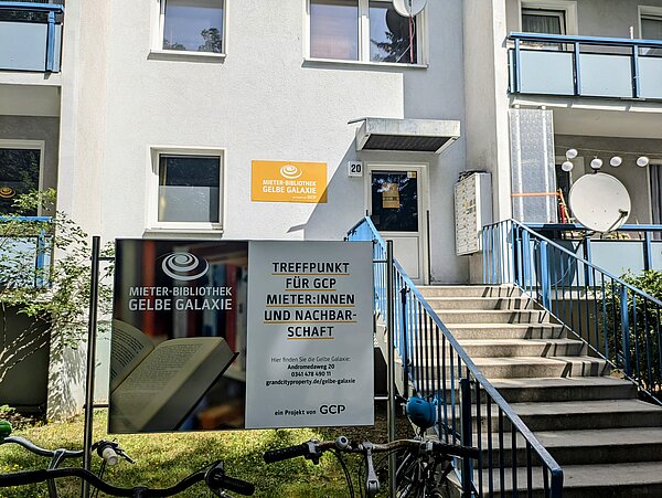Das Bild zeigt den Eingangsbereich der Mieter-Bibliothek Gelbe Galaxie. Es handelt sich um ein Mehrfamilienhaus mit weißer Fassade. Am Eingang, der über eine Treppe mit blauem Geländer erreichbar ist, befindet sich ein Schild mit der Aufschrift "Mieter-Bibliothek Gelbe Galaxie". Links im Bild sind Fahrräder zu sehen, die an einem Fahrradständer abgestellt sind. Am Gebäude selbst befinden sich Balkone mit blauen Geländern und eine Satellitenschüssel. Ein weiteres Schild auf dem Rasen vor dem Gebäude informiert über die Bibliothek als Treffpunkt für GCP-Mieter und die Nachbarschaft. 