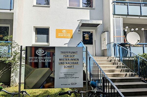 Das Bild zeigt den Eingangsbereich der Mieter-Bibliothek Gelbe Galaxie. Es handelt sich um ein Mehrfamilienhaus mit weißer Fassade. Am Eingang, der über eine Treppe mit blauem Geländer erreichbar ist, befindet sich ein Schild mit der Aufschrift "Mieter-Bibliothek Gelbe Galaxie". Links im Bild sind Fahrräder zu sehen, die an einem Fahrradständer abgestellt sind. Am Gebäude selbst befinden sich Balkone mit blauen Geländern und eine Satellitenschüssel. Ein weiteres Schild auf dem Rasen vor dem Gebäude informiert über die Bibliothek als Treffpunkt für GCP-Mieter und die Nachbarschaft. 