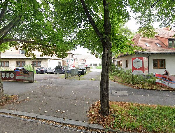 Das Bild zeigt das Gelände auf dem sich auch das Sportmuseums Leipzig befindet. Auf dem Gelände sind mehrere Gebäude mit hellen Fassaden und roten Ziegeldächern zu sehen. Links im Bild befindet sich eine bunte Wandgestaltung mit sportlichen Motiven, und auf der rechten Seite ist ein Gebäude mit mehreren Informationstafeln angebracht. Ein kleiner gepflasterter Weg führt auf das Gelände.