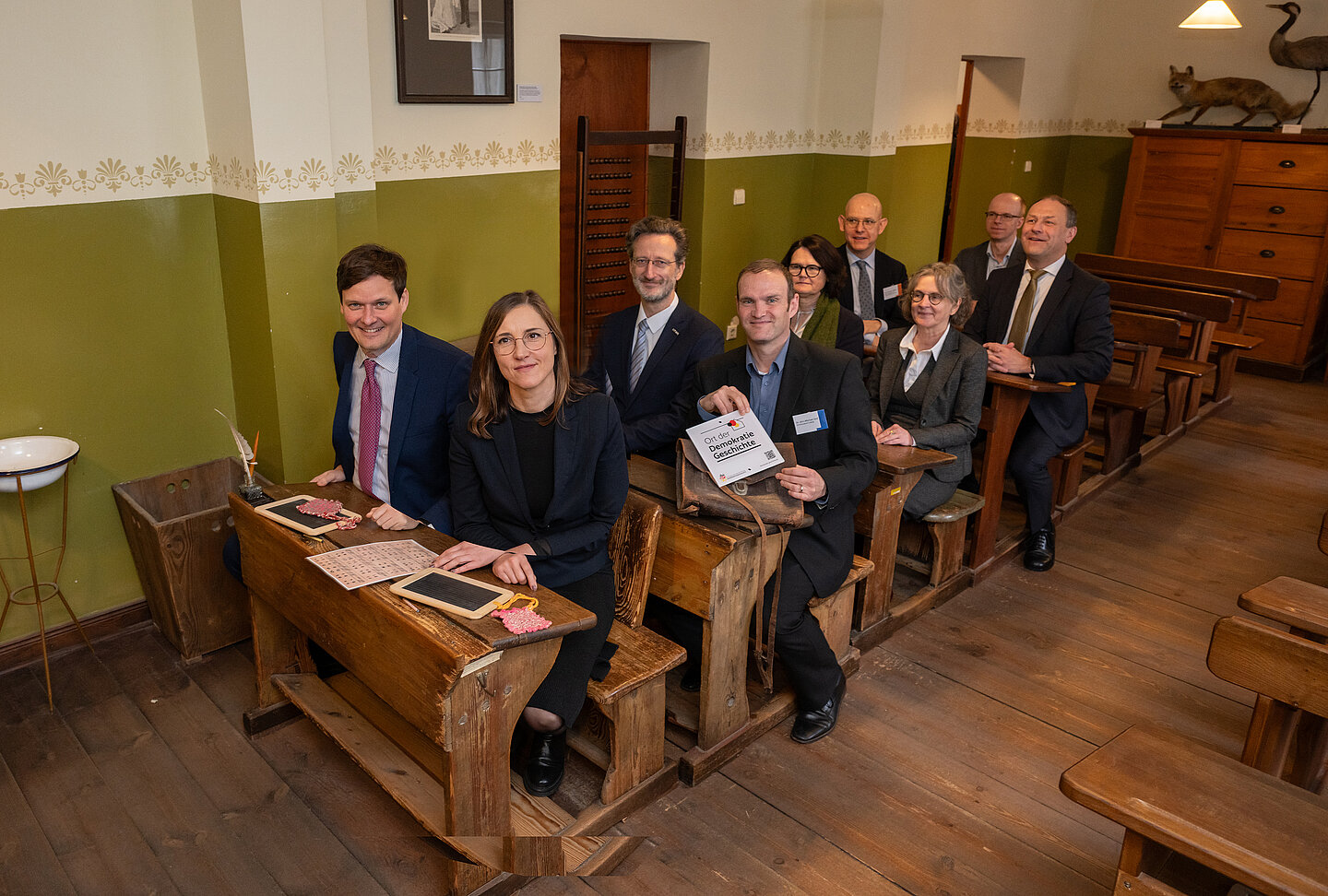 Erwachsene sitzen in alten Schulbänken in historischem Klassenzimmer
