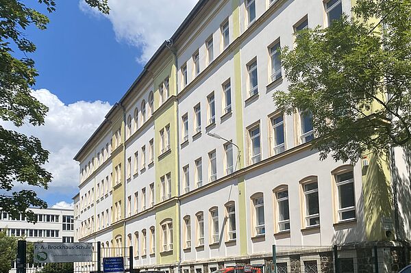 Das Bild zeigt das Friedrich-Arnold-Brockhaus-Gymnasium, ein großes, mehrstöckiges Schulgebäude. Die Fassade ist überwiegend weiß gestrichen, mit einigen grünlichen Akzenten an den Kanten und Fenstereinfassungen. Das Gebäude hat viele große Fenster, die gleichmäßig entlang der Wände verteilt sind. Ein Schild am Eingangstor weist auf den Namen der Schule hin: "F.-A.-Brockhaus-Schule – Gymnasium der Stadt Leipzig".