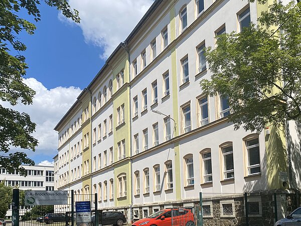 Das Bild zeigt das Friedrich-Arnold-Brockhaus-Gymnasium, ein großes, mehrstöckiges Schulgebäude. Die Fassade ist überwiegend weiß gestrichen, mit einigen grünlichen Akzenten an den Kanten und Fenstereinfassungen. Das Gebäude hat viele große Fenster, die gleichmäßig entlang der Wände verteilt sind. Ein Schild am Eingangstor weist auf den Namen der Schule hin: "F.-A.-Brockhaus-Schule – Gymnasium der Stadt Leipzig".