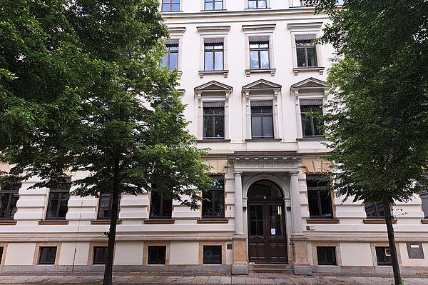Das Bild zeigt die historische Bibliothek des Thomanerchors in Leipzig, ein elegantes, mehrstöckiges Gebäude mit einer weißen Fassade. Charakteristische Merkmale sind die sorgfältig gestalteten Fenster mit steinernen Einfassungen und Giebeln, die über jedem Fenster angebracht sind. Der Haupteingang ist durch einen Bogen und eine Doppeltür hervorgehoben. Das Gebäude steht in einer ruhigen Straße, umgeben von reichlich Grün durch Bäume.