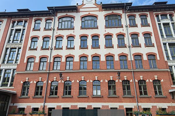 Das Bild zeigt die Johannes-Kepler-Schule, ein großes, historisches Gebäude mit einer Fassade aus roten Ziegeln und weißem Putz. Die vielen Fenster sind gleichmäßig angeordnet und haben dunkle Rahmen. Im oberen Teil des Gebäudes ist eine Uhr zu sehen. Vor dem Gebäude befinden sich einige Pflanzenkästen. Der Himmel ist blau mit einigen Wolken.