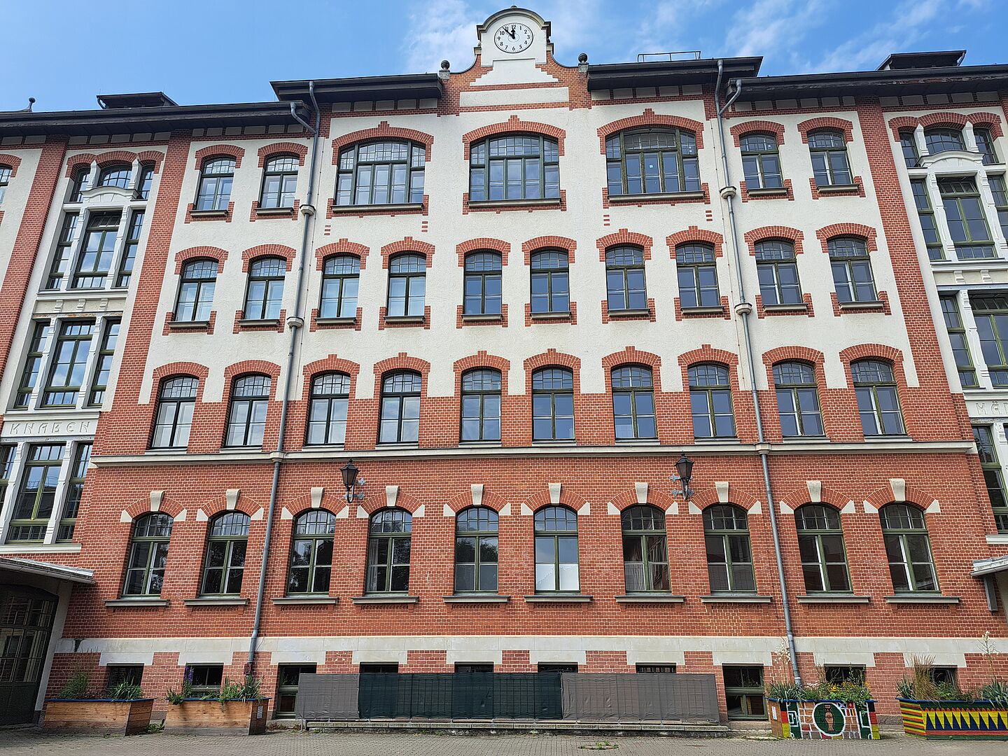 Das Bild zeigt die Johannes-Kepler-Schule, ein großes, historisches Gebäude mit einer Fassade aus roten Ziegeln und weißem Putz. Die vielen Fenster sind gleichmäßig angeordnet und haben dunkle Rahmen. Im oberen Teil des Gebäudes ist eine Uhr zu sehen. Vor dem Gebäude befinden sich einige Pflanzenkästen. Der Himmel ist blau mit einigen Wolken.