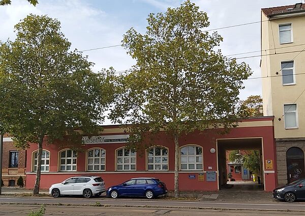 Das Bild zeigt eine langgezogene, einstöckige Fassade in einem rötlich-orangenen Farbton. Es handelt sich um ein Gebäude mit großen Fenstern, die gleichmäßig entlang der Front verteilt sind. Auf einem Schild an der Wand ist der Schriftzug "Vierseithof" zu erkennen. Auf der rechten Seite führt ein überdachter Durchgang zu einem Innenhof oder Hinterhof. 