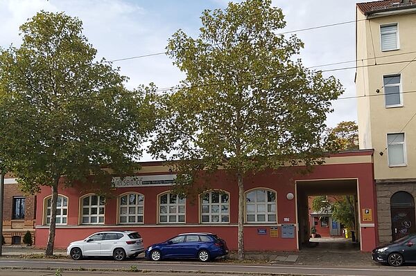 Das Bild zeigt eine langgezogene, einstöckige Fassade in einem rötlich-orangenen Farbton. Es handelt sich um ein Gebäude mit großen Fenstern, die gleichmäßig entlang der Front verteilt sind. Auf einem Schild an der Wand ist der Schriftzug "Vierseithof" zu erkennen. Auf der rechten Seite führt ein überdachter Durchgang zu einem Innenhof oder Hinterhof. 