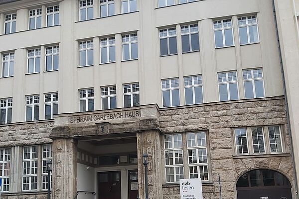Das Bild zeigt die Fassade des „Ephraim-Carlebach-Hauses“, einem mehrstöckigen Gebäude mit einer imposanten, steinernen Eingangspartie. Der Name des Hauses ist oberhalb des Eingangs in großen, blockartigen Buchstaben in den Stein eingraviert. Die untere Etage besteht aus massiven, steinernen Mauern, während die oberen Stockwerke in einem helleren, glatten Putz gehalten sind und durch viele Fenster mit weißem Rahmen gekennzeichnet sind. Vor dem Eingang befindet sich ein kleines Schild, auf dem "dzb lesen" steht, was für das "Deutsche Zentrum für barrierefreies Lesen" steht. Weiterhin bietet das Gebäude einen breiten Eingangsbereich mit Stufen, und es gibt eine Rampe, die für Barrierefreiheit sorgt.
