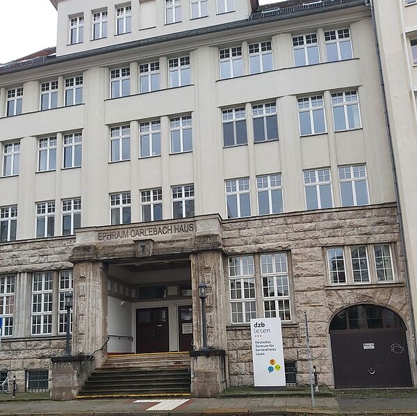 Das Bild zeigt die Fassade des „Ephraim-Carlebach-Hauses“, einem mehrstöckigen Gebäude mit einer imposanten, steinernen Eingangspartie. Der Name des Hauses ist oberhalb des Eingangs in großen, blockartigen Buchstaben in den Stein eingraviert. Die untere Etage besteht aus massiven, steinernen Mauern, während die oberen Stockwerke in einem helleren, glatten Putz gehalten sind und durch viele Fenster mit weißem Rahmen gekennzeichnet sind. Vor dem Eingang befindet sich ein kleines Schild, auf dem "dzb lesen" steht, was für das "Deutsche Zentrum für barrierefreies Lesen" steht. Weiterhin bietet das Gebäude einen breiten Eingangsbereich mit Stufen, und es gibt eine Rampe, die für Barrierefreiheit sorgt.
