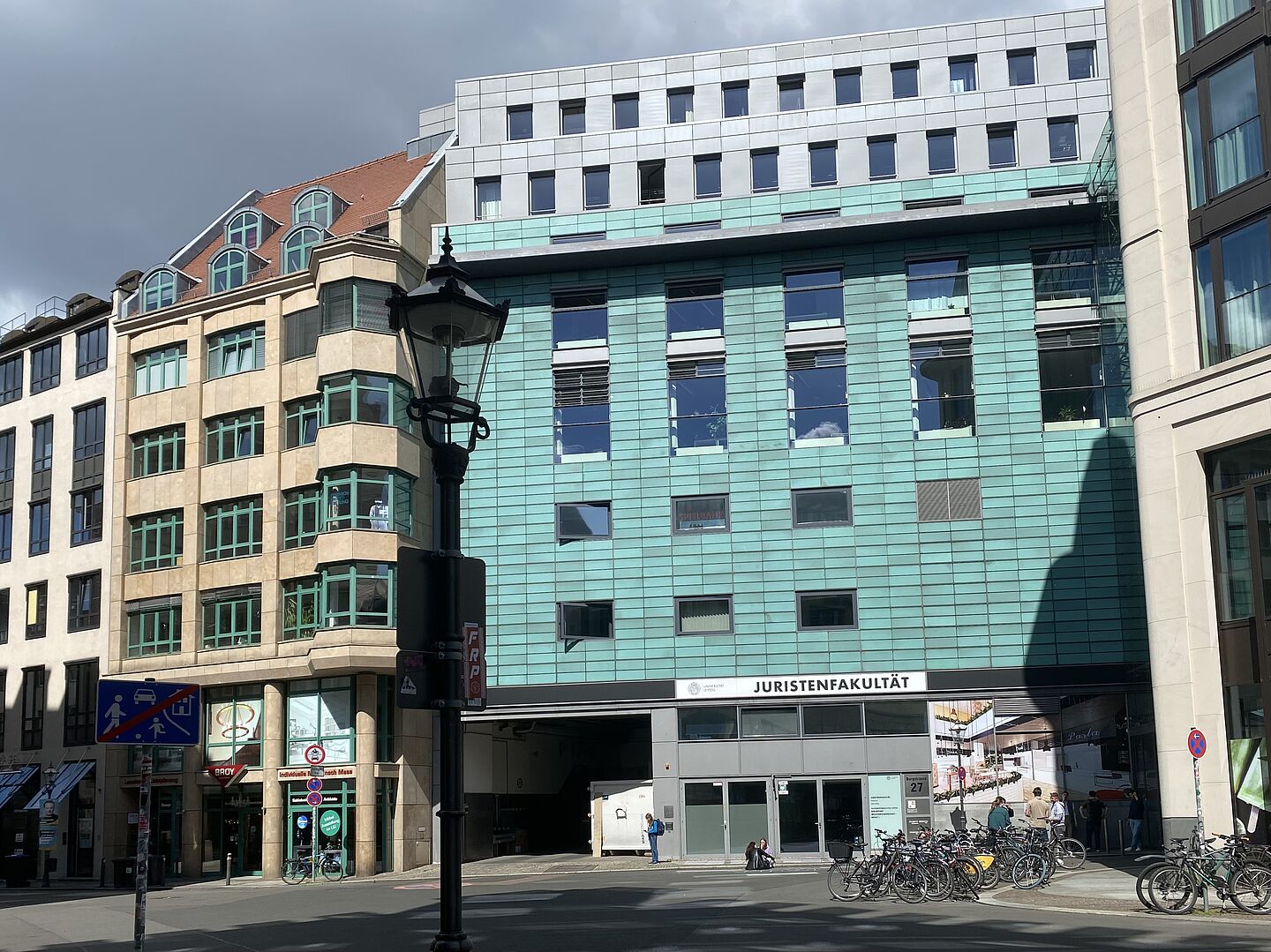 Das Bild zeigt ein Gebäude mit der Aufschrift „JURISTENFAKULTÄT". Das Gebäude hat eine moderne Gestaltung mit einer grünlichen Fassade und großen Fenstern. Links davon befindet sich ein weiteres Gebäude mit beiger Fassade und grünen Fensterrahmen. Im Vordergrund ist eine Straßenlaterne und mehrere Fahrräder sind nahe dem Eingang des Gebäudes abgestellt. 