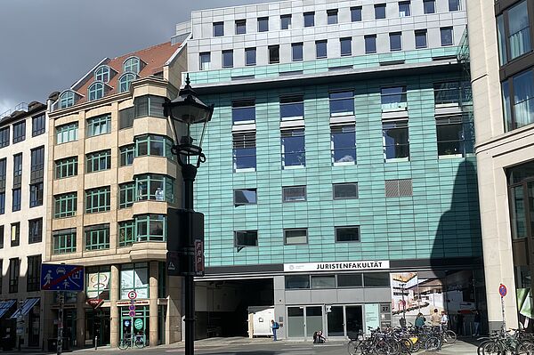 Das Bild zeigt ein Gebäude mit der Aufschrift „JURISTENFAKULTÄT". Das Gebäude hat eine moderne Gestaltung mit einer grünlichen Fassade und großen Fenstern. Links davon befindet sich ein weiteres Gebäude mit beiger Fassade und grünen Fensterrahmen. Im Vordergrund ist eine Straßenlaterne und mehrere Fahrräder sind nahe dem Eingang des Gebäudes abgestellt. 