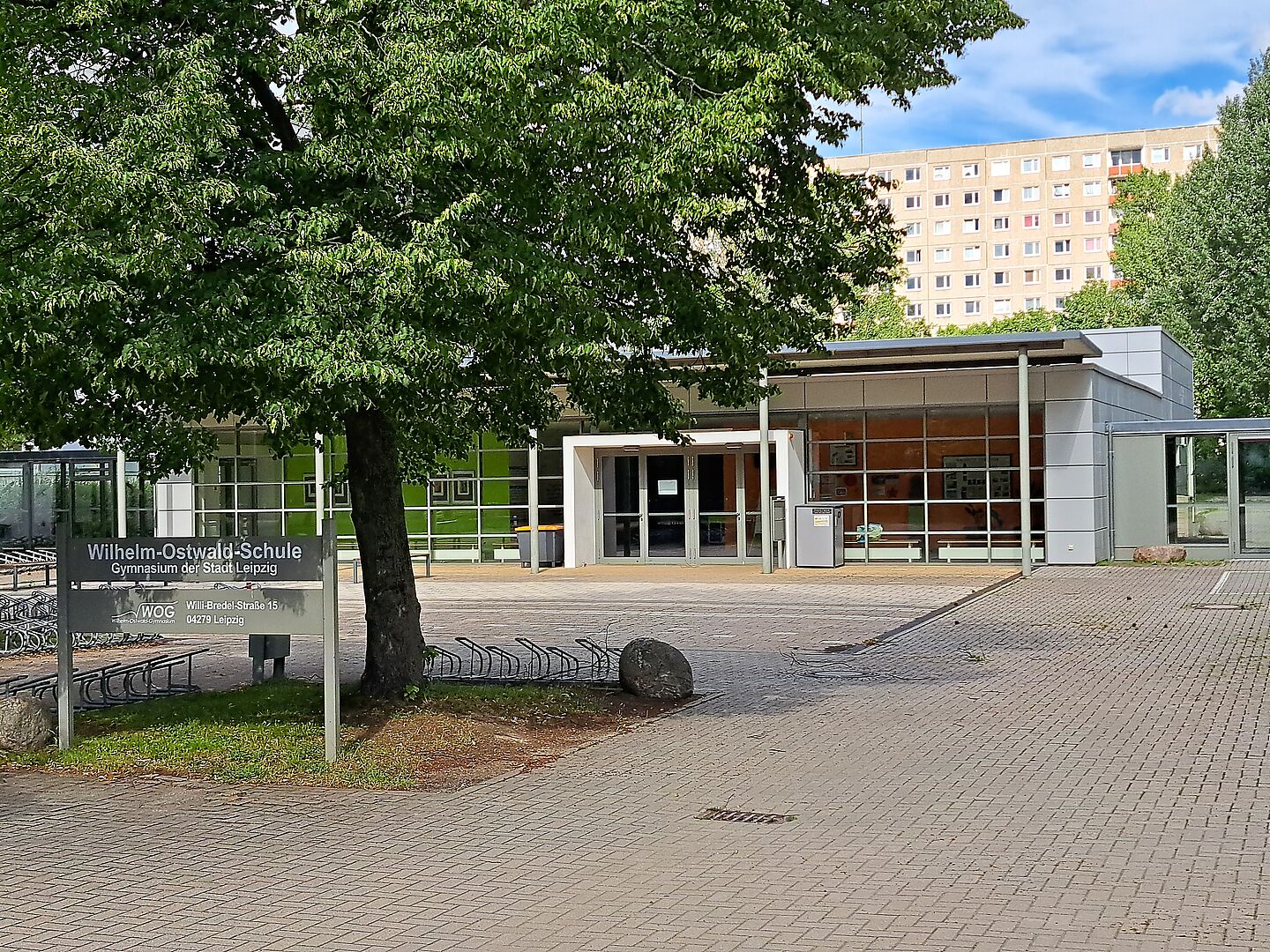 Das Bild zeigt das Schulgelände der Wilhelm-Ostwald-Schule. Man sieht ein flaches Schulgebäude und davor einen gepflasterten Platz mit einem Baum und einem Schild auf dem der Name der Schule steht.