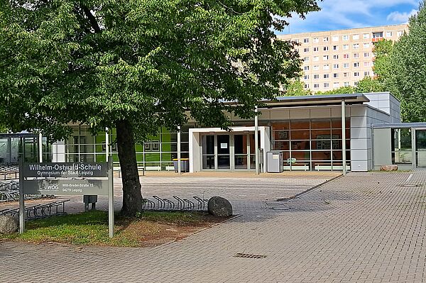 Das Bild zeigt das Schulgelände der Wilhelm-Ostwald-Schule. Man sieht ein flaches Schulgebäude und davor einen gepflasterten Platz mit einem Baum und einem Schild auf dem der Name der Schule steht.