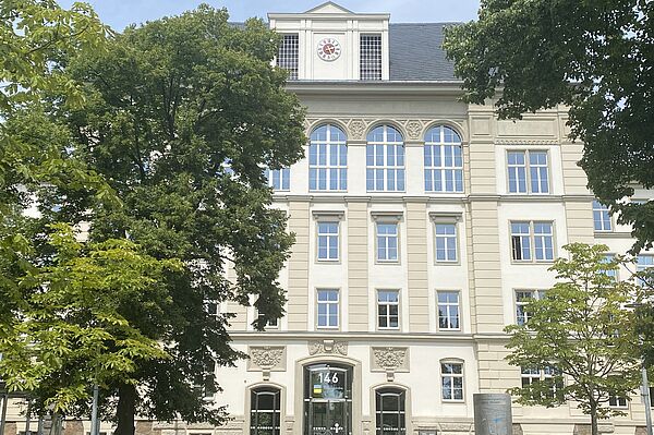 Ein mehrstöckiges Gebäude mit weißen Fassaden und mehrere große Fenster. Der Eingang besteht aus einer großen doppelten Glastür. Auf dem Dach ist eine Uhr zu sehen. Das Gebäude wird von großen Bäumen bedeckt.