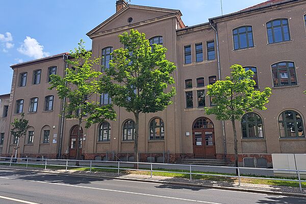 Das Bild zeigt ein mehrstöckiges, historisches Gebäude mit einer sandfarbenen Putzfassade und dunkelgrauen Fenstern. Vor dem Gebäude stehen mehrere junge Bäume. Eine asphaltierte Straße mit einem schmalen Gehweg und einer Absperrung verläuft im Vordergrund. Der Himmel ist blau und klar.