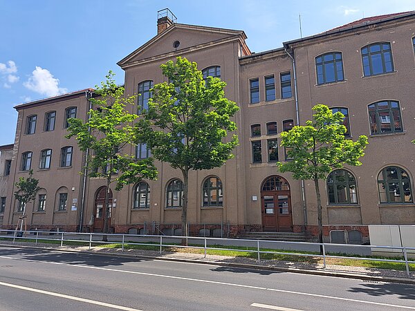 Das Bild zeigt ein mehrstöckiges, historisches Gebäude mit einer sandfarbenen Putzfassade und dunkelgrauen Fenstern. Vor dem Gebäude stehen mehrere junge Bäume. Eine asphaltierte Straße mit einem schmalen Gehweg und einer Absperrung verläuft im Vordergrund. Der Himmel ist blau und klar.