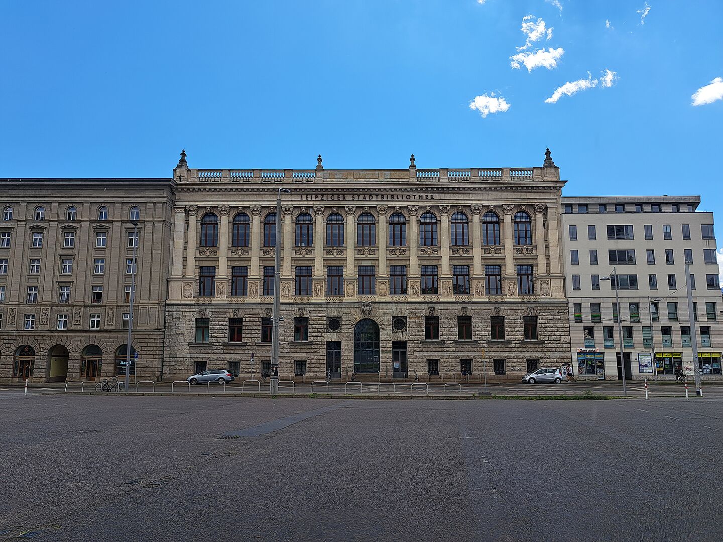 Das Bild zeigt die Stadtbibliothek Leipzig, ein großes, historisches Gebäude mit einer reich verzierten Fassade. Das Gebäude präsentiert eine Kombination aus robusten Steinsäulen und detailreichen Fensterumrandungen. An der Fassade sind Inschriften und ornamentale Details zu erkennen, die den klassischen Architekturstil unterstreichen.