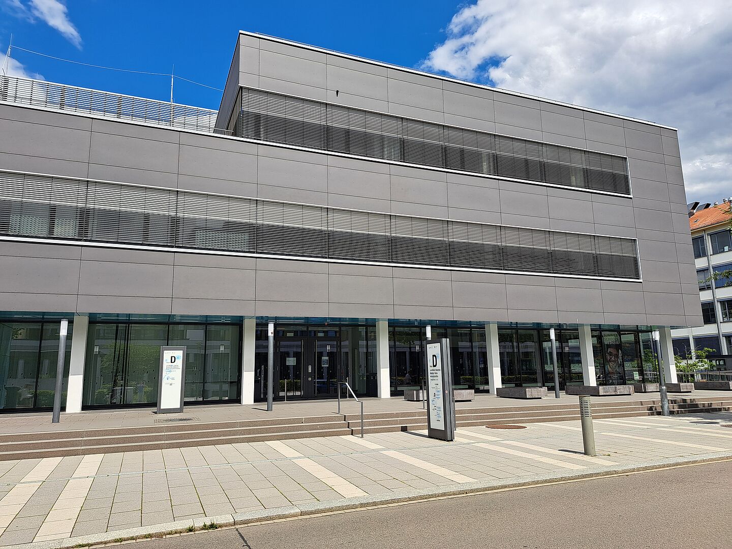 Das Bild zeigt die Universitätsbibliothek Leipzig, Bereich Medizin und Naturwissenschaften, ein modernes, mehrstöckiges Gebäude mit einer minimalistischen grauen Fassade. Die Vorderseite des Gebäudes ist mit großen Glasfenstern versehen, die eine klare Sicht in das Innere bieten. Vor dem Gebäude befindet sich ein gepflasterter Platz mit Zugängen zu mehreren Eingangstüren. Das Design ist schlicht und funktional.