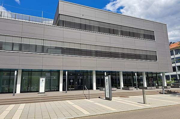 Das Bild zeigt die Universitätsbibliothek Leipzig, Bereich Medizin und Naturwissenschaften, ein modernes, mehrstöckiges Gebäude mit einer minimalistischen grauen Fassade. Die Vorderseite des Gebäudes ist mit großen Glasfenstern versehen, die eine klare Sicht in das Innere bieten. Vor dem Gebäude befindet sich ein gepflasterter Platz mit Zugängen zu mehreren Eingangstüren. Das Design ist schlicht und funktional.