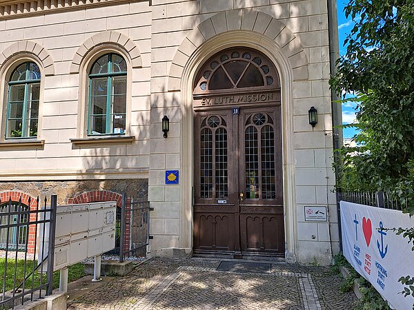 Das Bild zeigt den Eingang zum Evangelisch-Lutherischen Missionswerk e.V. mit einer großen Holztür und Bogenfenster. Die Fassade ist aus sandsteinfarbenem Material mit steinernen Details. Über der Tür ist der Name der Organisation eingraviert. Ein Wegweiser des Jakobswegs und ein Banner mit humanitären Symbolen sind neben dem Eingang sichtbar.