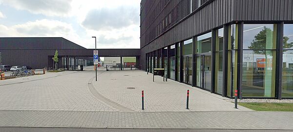 Dieses Foto zeigt ein modernes, architektonisch schlichtes Gebäude mit einer dunklen Fassade und großen Fensterflächen. Es handelt sich um einen Neubau mit einer klaren, geometrischen Gestaltung. Der Vorplatz ist mit hellen Pflastersteinen ausgelegt, und es gibt markierte Parkbereiche sowie eine klare Gehwegführung.