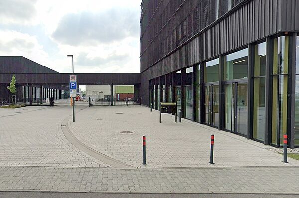 Dieses Foto zeigt ein modernes, architektonisch schlichtes Gebäude mit einer dunklen Fassade und großen Fensterflächen. Es handelt sich um einen Neubau mit einer klaren, geometrischen Gestaltung. Der Vorplatz ist mit hellen Pflastersteinen ausgelegt, und es gibt markierte Parkbereiche sowie eine klare Gehwegführung.