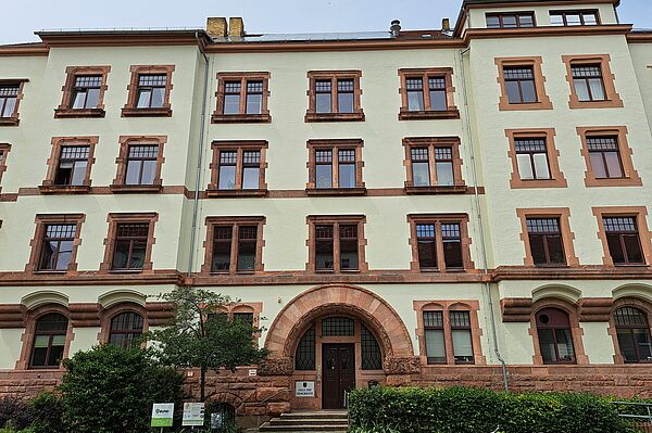  Das Bild zeigt das Haus der Demokratie. Es handelt sich um ein mehrstöckiges Gebäude mit einer cremefarbenen Fassade und rotbraunen Fensterrahmen sowie Verzierungen um die Fenster. Der Haupteingang ist von einem großen, rundbogigen Portal umgeben. Vor dem Gebäude befinden sich Fahrradständer und einige Büsche.