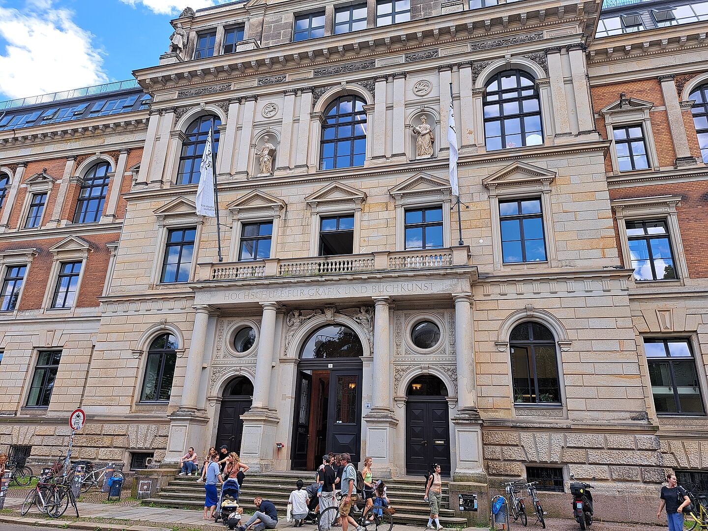  Das Bild zeigt die Hochschule für Grafik und Buchkunst Leipzig, ein elegantes Gebäude mit klassischer Architektur. Die Fassade kombiniert Elemente aus hellem und dunklem Stein und ist mit skulpturalen Verzierungen sowie Statuen geschmückt, die in Nischen platziert sind. Über dem zentralen Eingang, der durch eine doppelte Säulenstruktur hervorgehoben wird, steht der Name der Institution. Gruppen von Menschen sitzen auf den Stufen vor dem Gebäude.