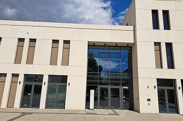 Das Bild zeigt die Universitätsbibliothek Leipzig für Erziehungs- und Sportwissenschaft, ein modernes Gebäude mit einer minimalistischen Fassade aus hellem Stein. Die Struktur ist geometrisch mit klaren Linien, großen Fensterflächen, die viel natürliches Licht ermöglichen, und rechteckigen Fenstern, die rhythmisch entlang der Fassade angeordnet sind. Der Haupteingang ist durch eine große Glasfront gekennzeichnet.