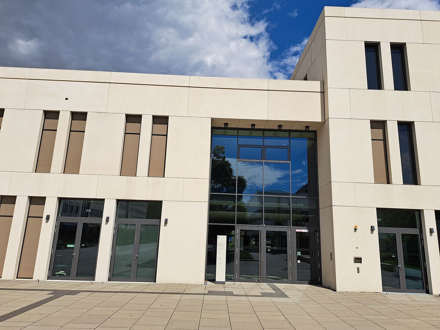 Das Bild zeigt die Universitätsbibliothek Leipzig für Erziehungs- und Sportwissenschaft, ein modernes Gebäude mit einer minimalistischen Fassade aus hellem Stein. Die Struktur ist geometrisch mit klaren Linien, großen Fensterflächen, die viel natürliches Licht ermöglichen, und rechteckigen Fenstern, die rhythmisch entlang der Fassade angeordnet sind. Der Haupteingang ist durch eine große Glasfront gekennzeichnet.