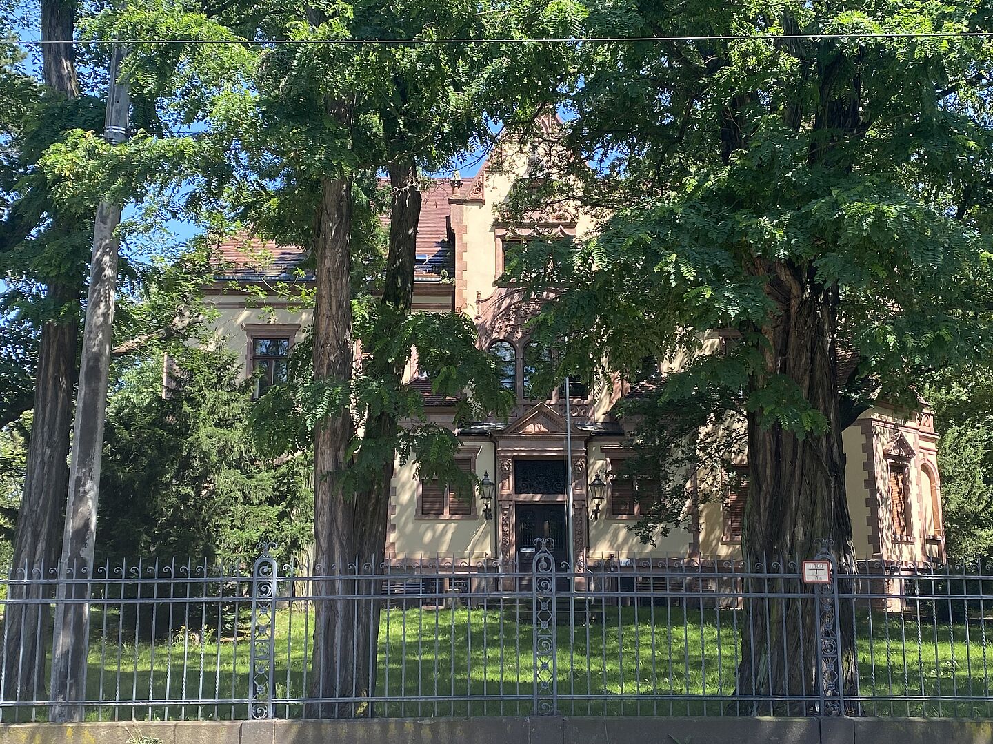 Das Bild zeigt ein historisches Gebäude, das teilweise von hohen Bäumen verdeckt wird. Die Fassade ist in einem warmen Beigeton gehalten, mit dekorativen Details in rötlichem Stein. Ein metallener Zaun umgibt das Gelände, und im Vordergrund ist eine gepflegte Grünfläche zu sehen. Das Gebäude besitzt mehrere Fenster und einen zentralen Eingang, der auf ein großes Eingangsportal hinweist.